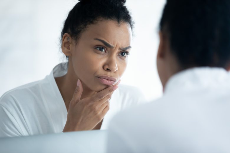 Dissatisfied worried young African woman scanning face reflection in mirror, checking skin for pimples, acne, wrinkles, treatment defects, mistakes. Dermatology, skincare problems, beauty care concept