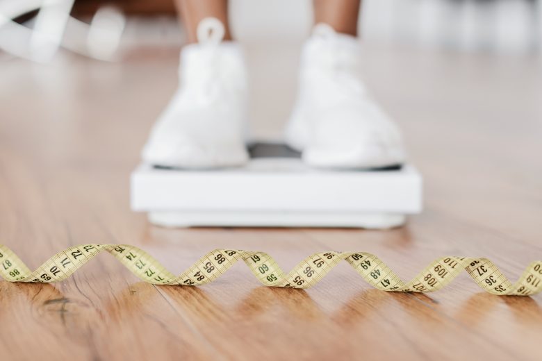 Weight Control Concept. Selective focus on yellow measuring tape on the floor and blurred view of african woman standing on scales, checking result of her slimming diet. Dieting, healthy living